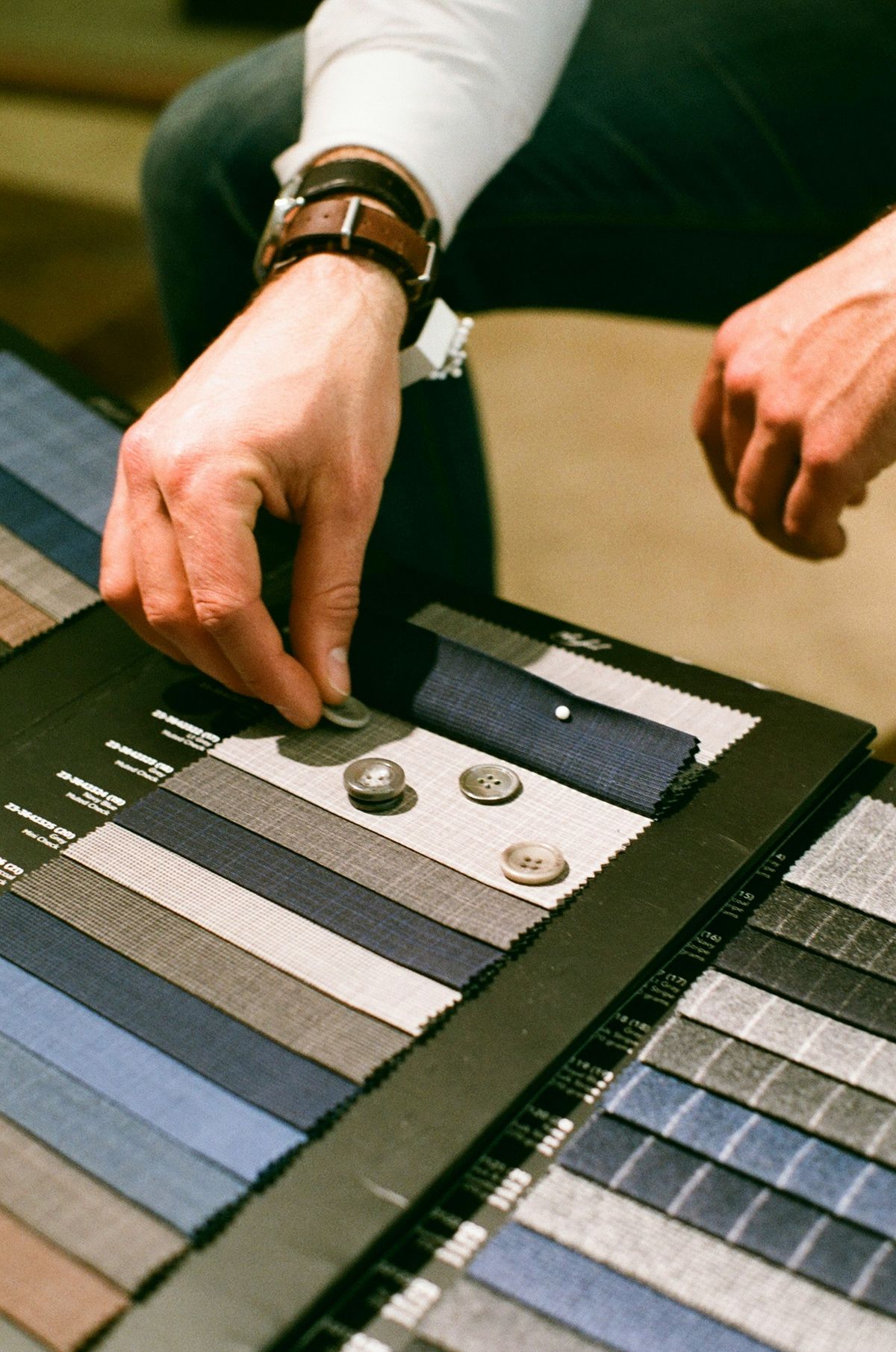 A hand placing a horn button on a swatch of grey worsted cloth, among books of navy, grey and blue suiting samples